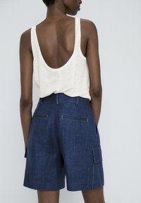 Personne portant un haut blanc sans manches avec un dos échancré et un short bleu foncé taille haute avec poche latérale, vue de dos.