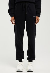 Černé tepláky s elastickým pasem a šňůrkou, nošené s bílými teniskami, s malým bílým logem na levé stehně.