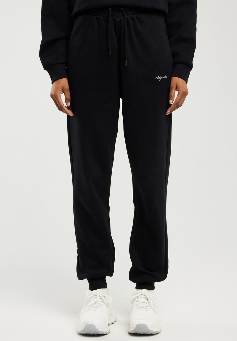 Černé tepláky s elastickým pasem a šňůrkou, nošené s bílými teniskami, s malým bílým logem na levé stehně.