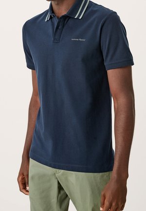 Mężczyzna ubrany w granatową koszulkę polo z paskowanym kołnierzykiem i napisem "letni smak", w połączeniu z jasnozielonymi spodniami.