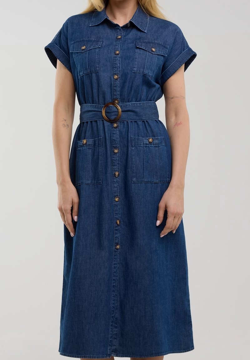Femme portant une robe midi en denim à boutons, manches courtes retroussées, poches poitrine et hanches, et une taille ceinturée avec une boucle ronde.