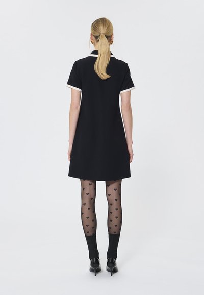 Vestido negro de mangas cortas con ribete blanco en el cuello, combinado con medias negras con patrón de corazones y zapatos de tacón negros. Silueta A-line sencilla.