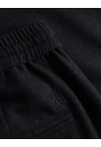 Un tissu en laine noir possède une ceinture froncée, avec une poche visible et une texture subtile. Logo 'II' brodé en accent sur le côté.