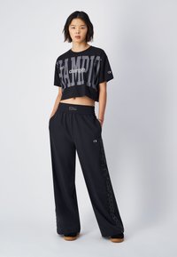 Zwarte cropped t-shirt met gedurfde "CHAMPION" graphics, gecombineerd met losse zwarte sportbroeken met elastische tailleband en logo-accenten aan de zijkant.