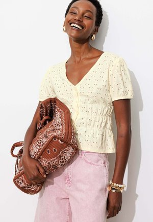 Femme souriante portant un chemisier crème à manches courtes et un pantalon rose, tenant un sac marron à motifs, avec des boucles d'oreilles en or et des bracelets mélangés.