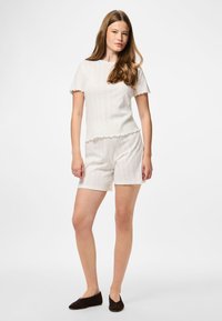 Mlada ženska z dolgimi lasmi, oblečena v off-white kratkosleeve pleteni top in ujemajoče kratke hlače, stoji na beli podlagi z črnimi čevlji.