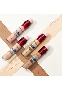 Quatre correcteurs multi-usages Maybelline présentent des tubes transparents avec des applicateurs en éponge rose. Les couleurs varient du beige clair au bronzage profond, avec les numéros 3, 5, 6 et 8 visibles.