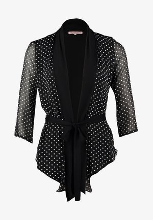 Kobieta ubrana w czarny przezroczysty kardigan w białe grochy na białym topie, czarne wąskie jeansy i czarne botki, trzymająca czarną kopertówkę.