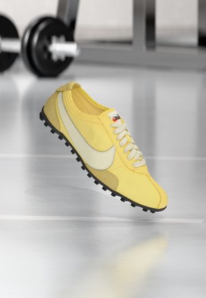 Žuta Nike tenisica za trčanje s bijelim zarezom i crnim đonom na reflektirajućem podu teretane, zamućene utege u pozadini.