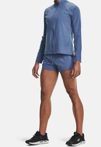 Person i blå långärmad zip-up-tröja, matchande shorts och svarta träningsskor, stående mot en enkel vit bakgrund.