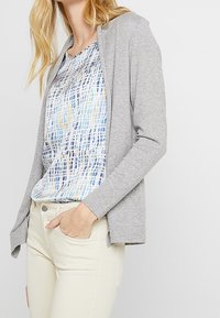 Cardigan grigio in tessuto morbido, design aperto davanti, maniche leggermente svasate, abbinato a un top a fantasia multicolore e pantaloni chiari.