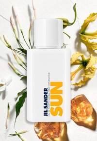 White Jill Sander Sun perfume bottle surrounded by yellow flowers, white buds, green leaves, and amber-colored stones on a textured surface.