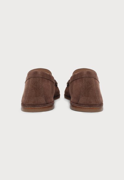 Paire de mocassins en daim marron à enfiler vus de dos sur un fond clair uni.