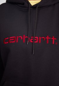 Black hoodie featuring prominent red embroidered "carhartt" logo, adjustable drawstring, and ribbed cuffs with a smooth cotton texture.