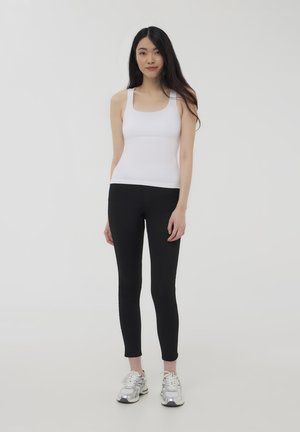 Top in cotone bianco senza maniche abbinato a leggings neri e scarpe da ginnastica argento, su uno sfondo bianco semplice.