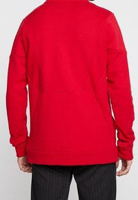 Czerwona bluza z gładką teksturą, z wysokim kołnierzem, długimi rękawami i detalami przeszyć na plecach, w zestawie z ciemnymi spodniami w paski.