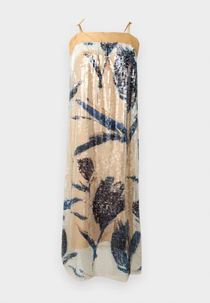 Femme portant une robe midi beige sans manches avec des motifs floraux bleus, des paillettes, des fines bretelles nouées aux épaules et des ballerines noires à bouts pointus.