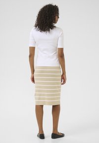 Un haut blanc ajusté associé à une jupe beige à mi-mollet ornée de rayures blanches horizontales. Des chaussures plates noires complètent la tenue.