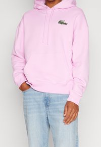 Felpa rosa in cotone con tasca a marsupio, cappuccio con coulisse e logo ricamato verde sul petto. Indossata con jeans denim chiari.