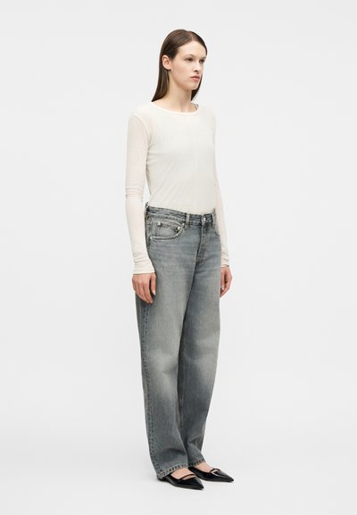 Γυναίκα που στέκεται φορώντας ψηλόμεσα ξεθωριασμένα γκρίζα τζιν, ημιδιαφανές λευκό μακρυμάνικο τοπ και μαύρες μυτερές μπαλαρίνες μπροστά σε λευκό απλό φόντο.