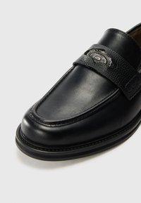 Black leather loafer featuring a textured strap with a decorative silver accent. Smooth finish, rounded toe, and sturdy sole.