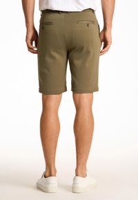 Vihreät oliivinväriset shortsit suoralla leikkauksella, takana kaksi nappipuolta ja käännetty helma; yhdistettynä valkoisiin lenkkareihin.