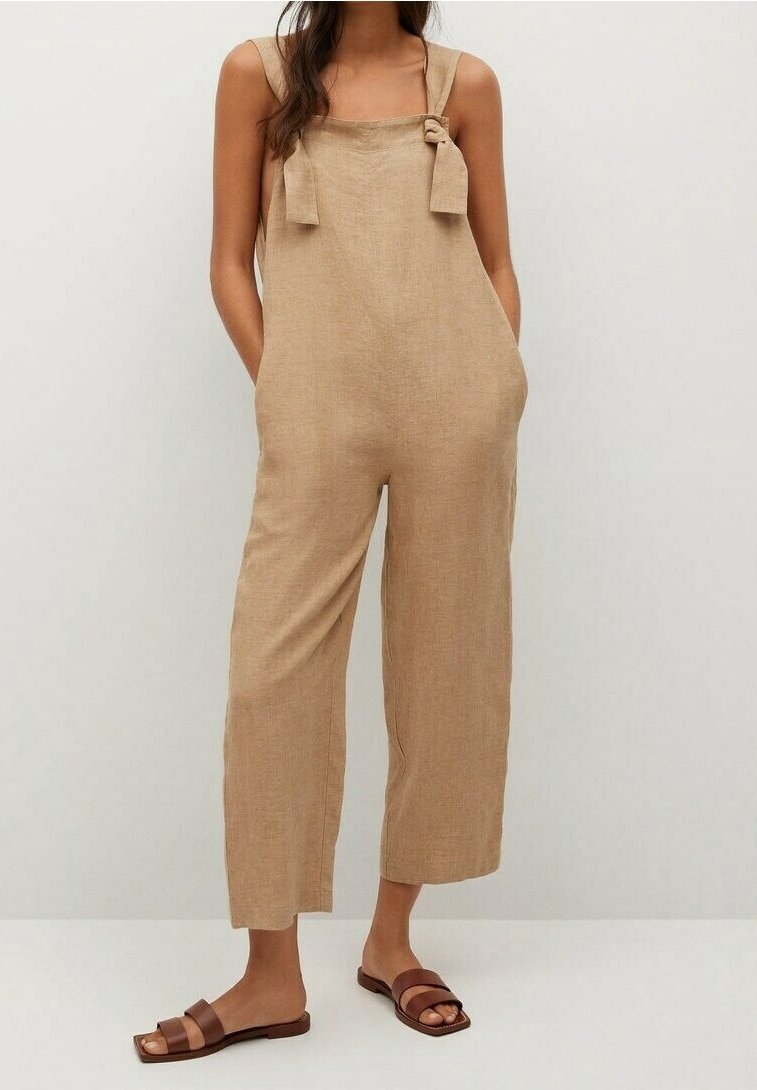 Femme portant une combinaison sans manches en lin beige avec des bretelles nouées aux épaules et des sandales plates marron, debout les mains dans les poches.