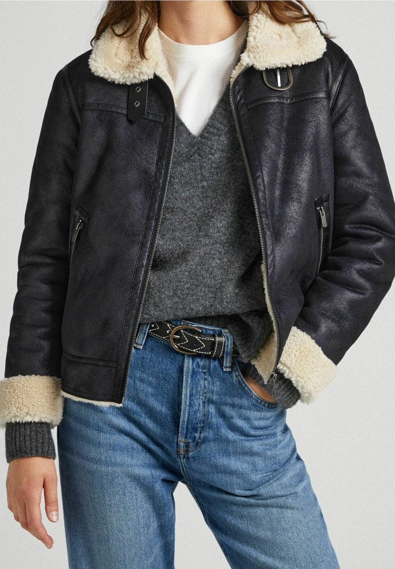 Veste en cuir noir avec un col en fausse fourrure beige, fermeture éclair à l'avant, poches latérales, associée à un pull gris et à un jean bleu.
