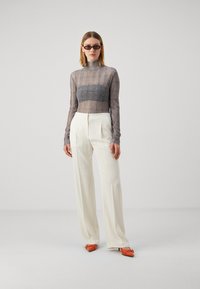 Top transparent à manches longues avec un motif à carreaux gris, associé à un pantalon blanc taille haute et des mules à talons oranges.