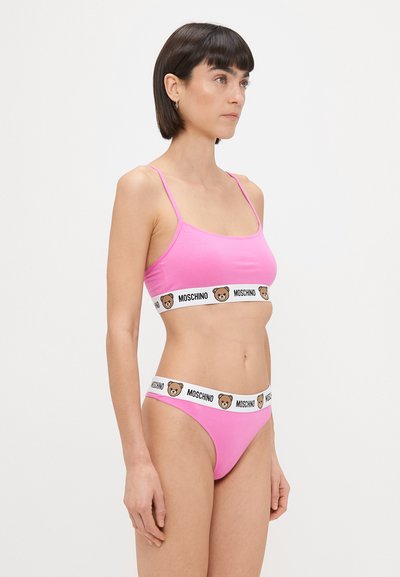 Roza bombažna bralette s tankimi naramnicami, ki ima pas z logotipom medvedka Moschino. Ujemajoči se roza bikini hlačke s podobnim logotipom na pasu.