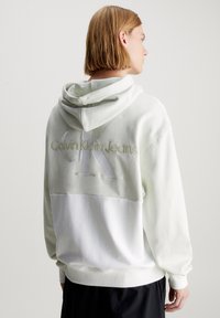 Sudadera verde claro con una sección inferior blanca, presenta un logo en relieve de "CK" y el texto "Calvin Klein Jeans" en la parte trasera. Ajuste suave y relajado.