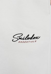 Biała tkanina o gładkiej fakturze, z haftem "Smilodox" w czerni i "ESSENTIALS" w jaśniejszym odcieniu poniżej.