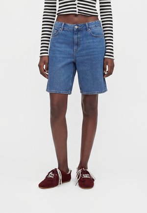 Person iført midt-lange blå denimshorts, en sort-hvid stribet cropped top og bordeaux sneakers med hvide snørebånd.