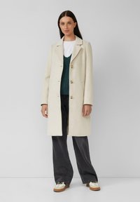 Manteau en laine beige clair avec de longues manches, col cranté et trois boutons. Porté par-dessus un pull en maille vert et un pantalon large gris.