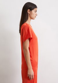 Red, short-sleeved top with a loose fit. Smooth texture and rounded neckline. Styled with matching pants, shown from the side.