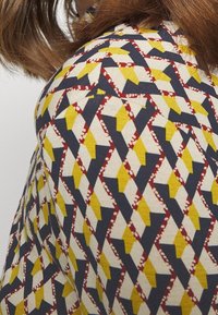 Stof med et geometrisk mønster der har gule, røde og marineblå diamanter og zigzag-mønstre på en lys baggrund, glat tekstur, nærsyn.