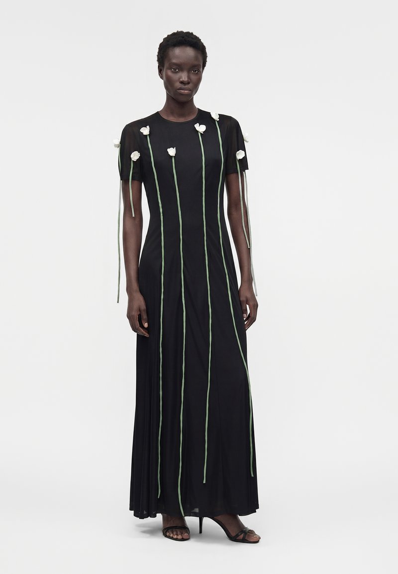 Femme portant une longue robe noire à manches courtes, décorée de grandes fleurs blanches et de tiges vertes qui descendent vers le bas.