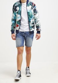 Chaqueta bomber colorida y estampada con un cuello de canalé azul marino, camiseta blanca, pantalones cortos de mezclilla y zapatillas altas negras.