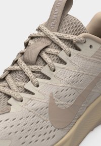 Close-up of beige Nike sneaker with textured fabric, detailed lacing, and visible "NIKE WILDPHORSE" text on tongue tab.