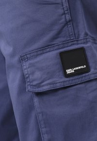 Tamnoplave cargo hlače od glatkog materijala, s bočnim džepom na kojem se nalazi crni kvadratni logotip s natpisom "KARL LAGERFELD JEANS."
