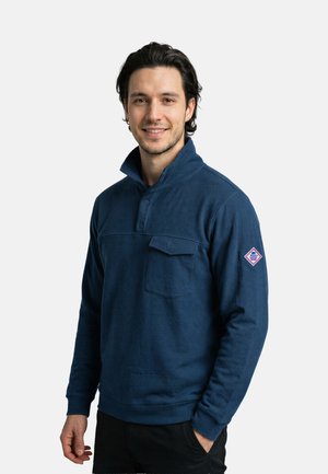 Junger Mann mit dunklem Haar, der einen marineblauen Pullover mit einer mit Knöpfen versehenen Brusttasche und einem Logo-Patch auf dem Ärmel trägt, lächelt in die Kamera.