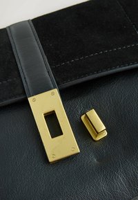 Borsa in pelle nera con pattina in camoscio, caratterizzata da una chiusura rettangolare in metallo dorato e un piccolo dettaglio dorato. Visibili dettagli della superficie testurizzata.