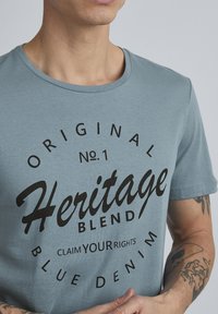 Svijetloplava pamučna majica s okruglim izrezom i crnim otisnutim tekstom: "Original Broj 1 Heritage Blend Zatraži Svoja Prava Plavi Denim."