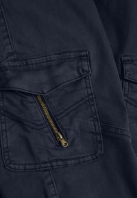 Mörk marinblå jacka med dubbla bröstfickor, varav en har ett guldzipper. Tyget har en slät textur och synliga sömnadsdetaljer.