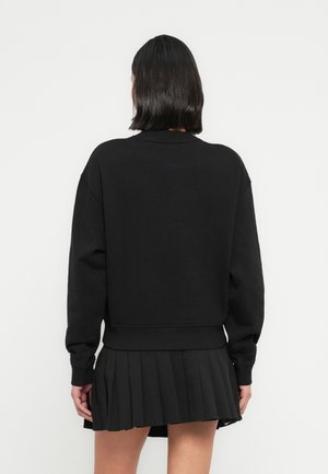 Person med skulderlangt sort hår iført en sort sweatshirt med lange ærmer og et sort plisseret mininederdel, vendt væk fra kameraet.