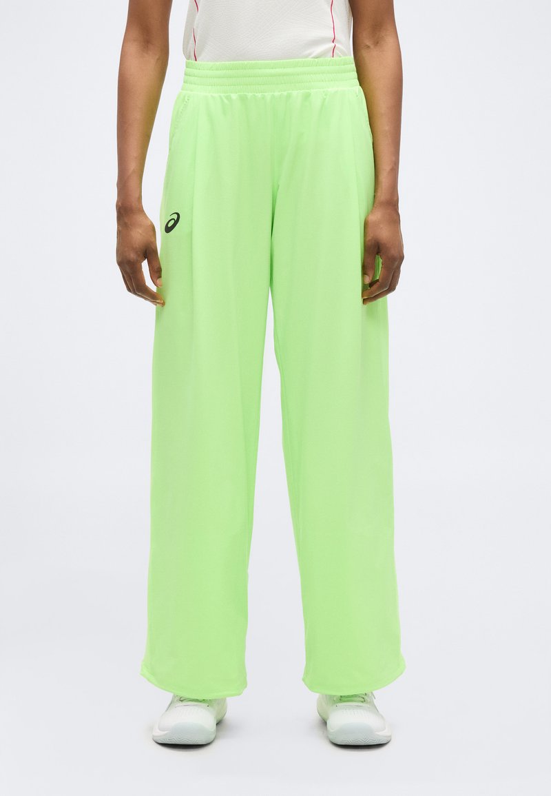 Άτομο που φοράει φαρδιά παντελόνια νέον πράσινου χρώματος με ελαστική μέση και λευκά αθλητικά παπούτσια, στεκόμενο μπροστά σε ένα απλό ανοιχτόφωτο φόντο.