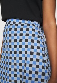 Person, die einen blau-schwarz karierten Rock mit einem seitlichen Reißverschluss und ein schwarzes Oberteil trägt, wobei Taille und Armbereich sichtbar sind.