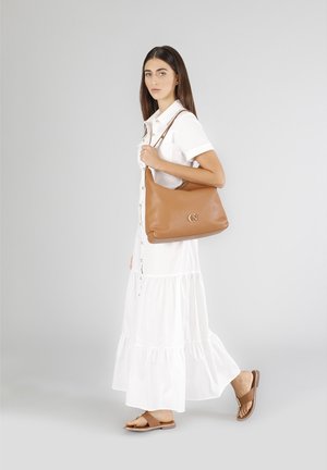 Brown leather shoulder bag with a smooth texture. Features a prominent logo accent and a single shoulder strap. Model wears a white shirt dress.
