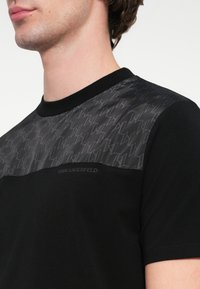 T-shirt preta com um painel cinza texturizado com um padrão geométrico nos ombros, decote canelado e logótipo bordado.