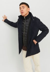 Abrigo de mezcla de lana azul marino sobre una chaqueta acolchada negra, con un cuello alto, cierre de botones y bolsillos laterales, combinado con pantalones crema.